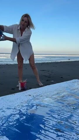 A blonde woman in a white open shirt and white bikini bottoms is on a beach, pouring blue paint from a bucket onto a large white canvas. She is barefoot and her legs are splattered with paint. There are several buckets of paint at her feet. The ocean is in the background with waves crashing. The sky is clear blue with a faint moon visible.