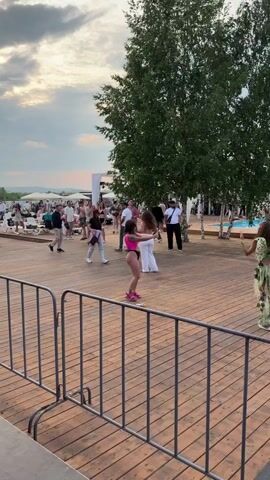 A group of people are on a wooden deck. Two women are dancing in the foreground. The woman on the left is wearing a bright pink swimsuit and white pants. The woman on the right is wearing a white halter top and white pants. There is a swimming pool in the background. The sky is cloudy.