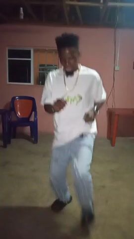 A young man is dancing enthusiastically indoors. He is wearing a white t-shirt with a stylized green graphic that appears to say "WAY" or similar, light blue jeans, and dark shoes. He also has a gold chain around his neck and a watch on his left wrist. The man is smiling and moving his arms and legs to the rhythm of the music, exhibiting an energetic and joyful demeanor.

The setting appears to be a simple room or a covered outdoor space, with light reddish-pink painted walls and a concrete floor. Exposed wooden beams are visible on the ceiling. In the background, there is a window with metal security bars. To the left, a blue plastic chair with an orange backrest is visible, and to the right, a small wooden table with an orange top. The lighting suggests it is evening or night. The overall scene indicates a casual environment, consistent with the location context of Iyara, Nigeria.