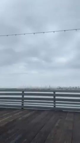 The video shows a drive across a wooden pier on an overcast day. Initially, the pier is empty except for a person walking in the distance. As the vehicle moves, trash cans, a fire extinguisher box, and a small building come into view. People can be seen walking on the pier near the building. The sky is cloudy, and the water is visible in the background. The video is shot from inside the vehicle, with a view of the pier and the surrounding environment.