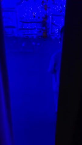 A young boy wearing a white t-shirt and shorts stands in a room illuminated by blue light. The walls and floor are reflective, creating distorted images and patterns of light. In one section of the video, a person walks through the room, their silhouette visible against the bright blue reflections. The room appears to be an art installation or a space with interactive lighting and reflective surfaces, possibly an exhibition or a mirrored maze.