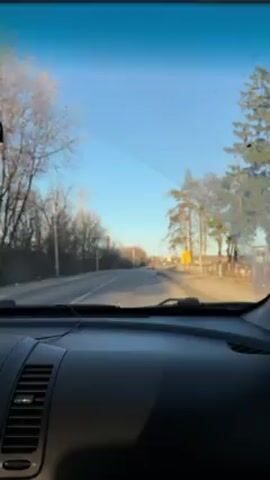The video shows a view from inside a car, looking out through the windshield while driving on a paved road. The sky is clear and blue, and the sun appears to be shining. Trees line the side of the road, mostly bare of leaves. There is a yellow road sign visible on the right side of the road. Another car is seen driving in the opposite direction, approaching the camera. The dashboard of the car taking the video is dark.