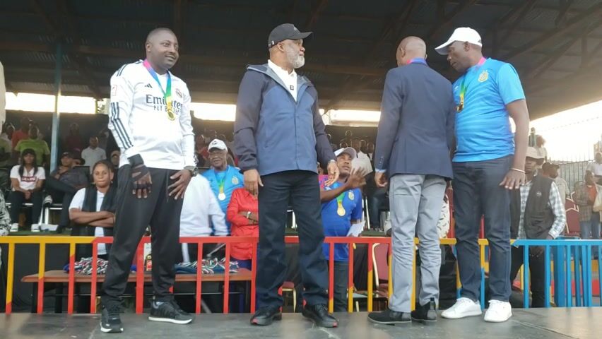 Here is a concise description of the video:

Four men are standing on a stage in front of a crowd. All four men are wearing medals around their necks, presumably indicating they are being honored for something. The man on the left is wearing a white and black soccer jersey. The second man from the left is wearing a navy blue jacket. The third man is wearing a blazer and light gray pants. The man on the right is wearing a blue soccer jersey. The man on the left is talking to the man in the navy blue jacket, possibly explaining something about the medal or event. The background shows a crowd of people seated in stadium-style seating. The stage is bordered by a colorful railing. The scene suggests a celebratory atmosphere, possibly after a sporting event or ceremony.