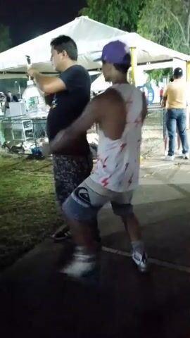 Two men are standing near a white tent in an outdoor area at night. One man in a black shirt and printed pants is holding a phone and filming the other man. The second man is wearing a white tank top with red lightning bolts, patterned shorts, a purple baseball cap, and sneakers. He moves slightly, seemingly posing for the camera. The area is somewhat littered with debris, and other people can be seen in the background. A fence runs along the edge of the walkway where the men are standing.