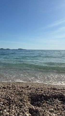 The video shows a rocky beach on a sunny day. The sea is calm and turquoise in color, with gentle waves lapping at the shore. In the distance, there are a few small islands visible on the horizon. The sky is a clear blue with few clouds. No people are visible in the shot.