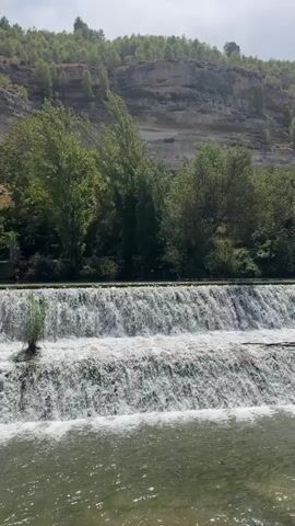 A video shows a cascading waterfall in a natural setting. The waterfall is made up of multiple tiers, with water flowing over each level and creating white foam. The foreground of the video features the river water below the waterfall, which is a murky green color and appears to be flowing. In the background, there are lush green trees and a rocky cliff face covered in vegetation. The sky is overcast, suggesting a cloudy day. There are no people or man-made objects clearly visible in the frame, apart from what appears to be a constructed element at the top of the waterfall, possibly a dam or weir. The overall impression is one of a serene natural landscape with the dynamic element of flowing water.