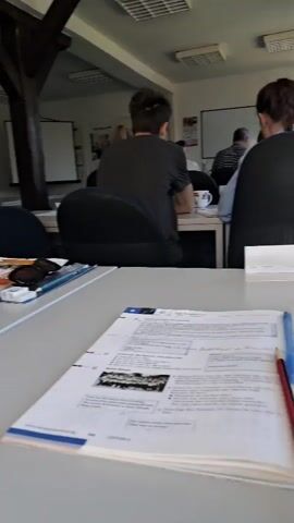 The video pans across a classroom or lecture hall with several people seated at tables, facing forward. The camera is positioned low, suggesting it might be on a table or desk. In the foreground, a book with text and images is open, with a blue pen and a red pencil lying on top of it. To the left, another person's arm is visible, holding a pen and making notes in a notebook. The room has fluorescent lighting on the ceiling and a large wooden beam running diagonally across it. There are projection screens visible at the front of the room, and windows along one side. People are dressed casually, and some appear to be engaged in writing or listening. The overall impression is of a learning or meeting environment.