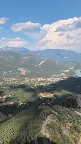 A drone shot pans over a vast mountainous landscape dotted with turquoise lakes. A small town is visible nestled between the hills and one of the lakes. The sky is blue with fluffy white clouds casting shadows on the terrain below. Lush green trees and rocky outcrops cover the foreground.
