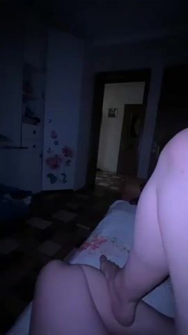 The image depicts a dimly lit indoor scene, likely a bedroom. The foreground features a person's legs and feet resting on a floral-patterned surface, possibly a bed. The legs are bare, and the feet are positioned near the center of the frame.

In the background, a closet with floral decals is visible to the left. Further back, an open doorway reveals a hallway with tiled flooring and another door. A framed picture hangs on the wall in the hallway. The overall lighting suggests it is nighttime or the room is kept dark. The setting appears to be a residential interior. No specific activity is clearly discernible, beyond the person resting. No text is visible in the image. The location context of Zhigalovo, Russia, is provided but not visually identifiable from the image itself.