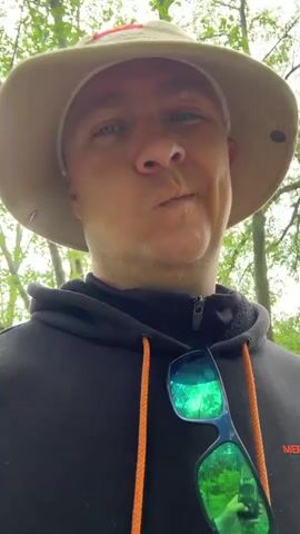 The video shows a close-up shot of a man wearing a tan safari hat with a Vietnamese flag emblem, a black Merrell hoodie with orange drawstring, and reflective green sunglasses hanging from his neck. He is standing outdoors in a natural setting, possibly a park or forest, with trees visible in the background and what appears to be a body of water partially visible through the trees. The man looks directly at the camera with a slight smirk or pursed lips expression. He is moving slightly, suggesting the video is being taken while walking.