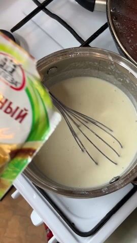 In a close-up shot, a person is pouring ground nutmeg from a green and white package into a stainless steel saucepan filled with a white liquid. The saucepan sits on a white gas stove. A metal whisk is submerged in the liquid. A second, covered saucepan is visible in the background. The package of nutmeg is labeled in Russian.