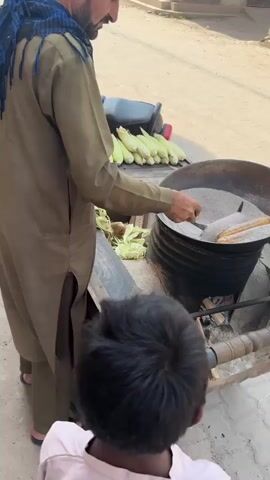 A man in a tan shalwar kameez and blue scarf is using a metal spatula to rotate corn on the cob in a large wok-like pot filled with grey sand. The pot sits on a brick and metal base over a fire. A small boy watches him from the front of the scene. The man stands next to a stack of green corn cobs on a metal surface. A parked motorcycle is in the background on a street with buildings on either side.
