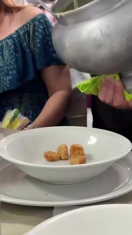 A person in a blue dress is seated in the periphery of the video. A metal pot is being held by a person obscured from view, who is using a green cloth to support the pot. A ladle is dipping into the pot, pouring a pale green soup over several croutons in a white bowl. The bowl sits on a white plate on a light surface.