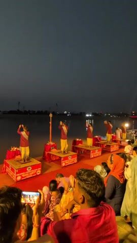 The video shows a group of five individuals dressed in red and yellow clothing performing a ritual by a body of water at night. They are standing on raised platforms adorned with red fabric, each with a lit lamp and a trident-like object. The background features a dark sky with distant lights, and a crowd of people are seated in the foreground, some holding phones to record the event. The overall atmosphere appears to be a religious or cultural ceremony.