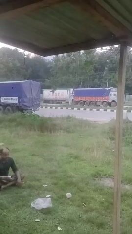 A video recording displays a roadside scene with several trucks. In the foreground, a man sits cross-legged on grassy ground under a covered structure, possibly a shed or overhang. In the background, multiple trucks are visible on a road that runs parallel to the grassy area. The trucks appear to be in transit, some covered with blue tarpaulin. The environment suggests an outdoor, possibly rural or semi-urban setting with trees and vegetation. The lighting indicates it is daytime.