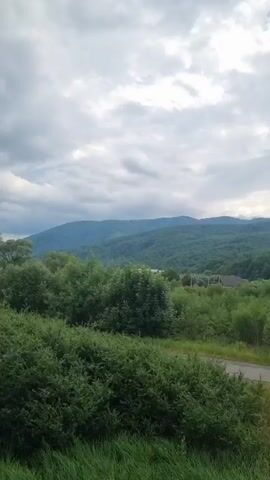 The video shows a scenic landscape with rolling green hills and mountains in the background. The sky is cloudy, with patches of sunlight breaking through. In the foreground, there is a road and a grassy field. There are also some trees and bushes scattered throughout the scene. A small village or collection of houses can be seen nestled in the valley between the hills. The overall impression is one of peaceful rural life.