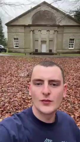 A young man with short hair, wearing a blue long-sleeved shirt with white flower designs, is taking a selfie video while walking on a path covered with fallen leaves. Behind him is a stone building with a classical facade. The sky appears overcast, and there are trees visible in the background. He is looking directly at the camera.