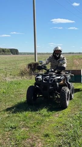 A man in a helmet and camouflage jacket rides a four-wheeled ATV with a small green trailer through a grassy field on a sunny day. The ATV is dark colored with “JAEGER” displayed on its side. The field has some dirt tracks and is surrounded by trees. There is a light pole in the background. The sky is blue with scattered clouds.