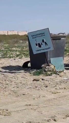 The video shows an outdoor scene with several large, dark-colored garbage bins. In the foreground, there is a sandy area with some scattered debris. A sign is propped up against one of the bins, displaying an image of people and text in a foreign language. To the right of the bins, there appears to be a rusted barrel with some contents inside, and a piece of blue glass or plastic is lying on the ground. Further down, a small, black toy jeep is parked next to one of the bins, with some white items inside it. The environment appears somewhat desolate with sparse vegetation and distant buildings visible in the background. The overall impression is one of an unkempt public space.