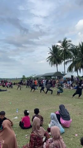 A wide shot shows an outdoor tug-of-war competition in progress under a cloudy sky with palm trees in the background. A group of people are seated in the foreground, facing away from the camera and towards the competition. In the middle ground, two teams of men are pulling on a rope. One team is wearing dark clothing, while the other team is wearing a mix of dark and green clothing. There are spectators standing and sitting behind the competitors. Tents and a stage are visible in the far background. The ground is covered in grass and scattered with plastic bottles. The overall scene suggests a community event or festival.