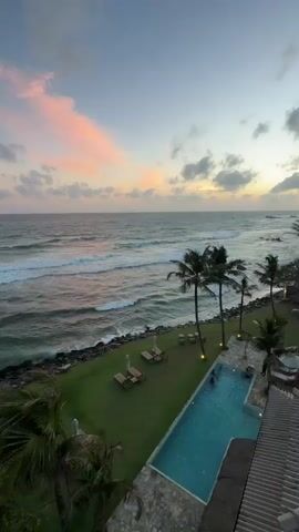 The video pans over a scenic coastal resort at sunset. The ocean is visible in the upper left of the frame, with waves breaking against a rocky shore. A strip of green grass separates the shore from a swimming pool in the lower right. Several lounge chairs are scattered on the lawn. Palm trees line the edge of the pool. Two people are swimming in the pool. The sky is a mix of blue and pink hues, with scattered clouds. The overall impression is one of relaxation and natural beauty.