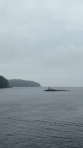 The video shows a wide shot of a body of water under an overcast sky. In the distance, a forested island is visible on the left. To the right of the island, on a small rocky outcrop, stands a white lighthouse. The water in the foreground has small ripples, indicating a light breeze. The overall mood is calm and somewhat somber due to the grey weather.