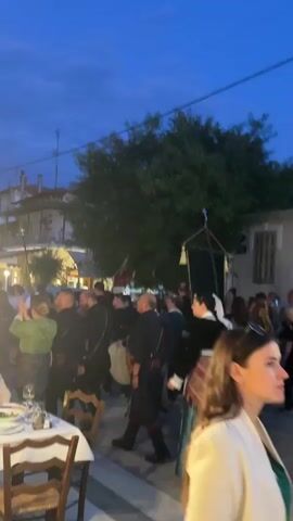 The video shows a street scene in what appears to be a town or village. The sky is dark, suggesting it is evening or dusk. Several people are visible, some dressed in traditional clothing. In the foreground, there is a table with chairs, hinting at an outdoor dining area. Further back, a crowd is gathered, with some individuals holding banners. A prominent sign for "ANDREAS RESTAURANT" is visible on a building. People are walking around, and there are lights illuminating the street and businesses. The overall atmosphere suggests a cultural event or a lively gathering.