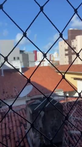 A security net is in the foreground, obscuring a view of a city skyline. The net is black and has a diamond pattern. Beyond the net, the sky is blue with some clouds. The middle ground is filled with buildings and rooftops. Some of the buildings are tall and modern, while others appear older and are made of brick. The rooftops are predominantly made of orange terracotta tiles. In the foreground, near the bottom of the frame, there are several structures on the rooftops, including what appears to be a satellite dish and a chimney. The overall impression is of an urban environment on a sunny day.