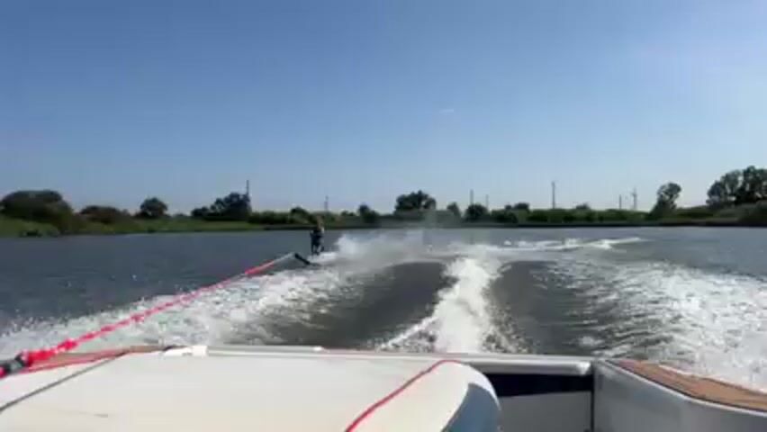 The view from a moving boat shows a person water skiing on a lake. The skier is being pulled by the boat, and they are creating a spray of water as they move across the lake. The lake is surrounded by trees and fields, and the sky is blue. The boat's wake trails behind.