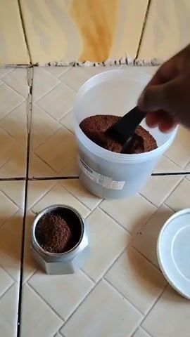 A hand uses a small black measuring spoon to fill the silver bottom half of a moka pot with ground coffee. The coffee is being scooped from a large translucent plastic container with a label on the side. The white plastic lid to the container is on the tile floor to the right of the container. The tile floor is a light cream color.