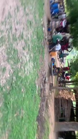 The video shows an outdoor scene with a group of people gathered. The camera appears to be moving, causing a blurry effect. In the foreground, there is grass and dirt. Further back, there are people sitting on chairs and what appears to be a gathering or event. Some individuals are visible in the frame, and there are trees and foliage in the background. The orientation of the video is sideways, with the bottom of the frame appearing on the left.