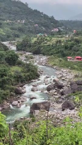 A video shows a wide river flowing through a valley. The river has many rocks and some rapids. The banks of the river are covered in green trees and vegetation. In the background, there are hills with houses built on them. The sky is overcast.