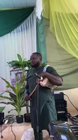 A man is standing indoors, holding a small drum and a curved drumstick. He is looking at the camera and gives a peace sign. He is wearing a green tunic and pants. Behind him is a green and yellow curtain, and to his left are two potted plants. There is sound equipment to his right.