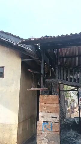 A video shows a structure that appears to have been damaged by fire. The structure is largely blackened and charred, with visible damage to the roof and walls. A person is using a hose to spray water onto the structure, presumably to extinguish any remaining embers or prevent further damage. Other people are visible in the background, observing the scene. The environment appears to be outdoors, with what looks like trees and other buildings in the distance. The sky is visible and appears overcast or hazy. The overall scene suggests a recent fire incident and an ongoing effort to manage the aftermath.