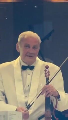 A video begins with a close-up shot of an older Caucasian man wearing a white tuxedo and black bow tie, holding a violin and bow. He is looking directly at the camera with a neutral expression. The background is dark and out of focus, with some blurred lights. The camera then pulls back slightly to reveal more of his torso and the surrounding environment. He is seated at a table with a menu in front of him. Behind him, a large banner or backdrop is visible, featuring a ship graphic and text in a foreign language. The man gestures with his free hand as he speaks. The camera continues to pull back, showing a dimly lit room with tables and chairs, suggesting a dining or performance venue. The man remains the central focus, continuing to speak and gesture. The video ends with a wider shot of the room, with the man still visible at the table, holding his violin.