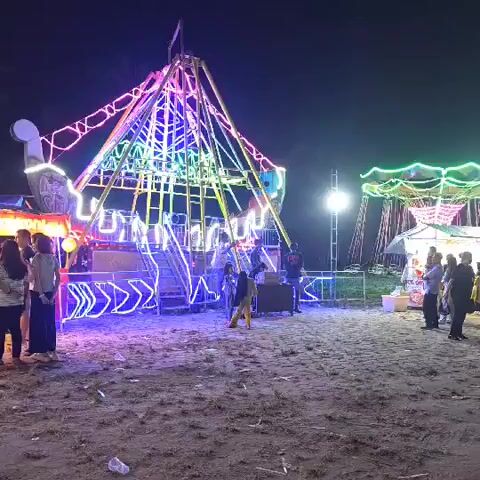 The video shows a night time scene at an outdoor amusement park or fair. Several rides are visible, including a tall swing ride with colorful lights, a carousel with lights, and a smaller ride that appears to be a bouncy castle or similar attraction. People are gathered around the rides, and some appear to be waiting in line or interacting with vendors. The ground is sandy, suggesting the location might be near a beach. The lighting from the rides illuminates the scene, creating a festive atmosphere.