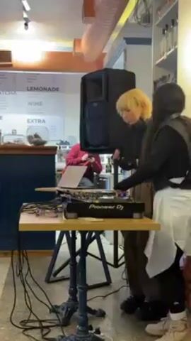Two women are operating a Pioneer DJ mixing console on a small table, situated in what appears to be a cafe or restaurant. A large black speaker stands beside them. One of the women has short blonde hair and is wearing a black top and brown pants. The other has long dark hair and is wearing a black top with a white apron or garment. The table is covered in cables, and a laptop is open near the mixer. A man in a red shirt can be seen using his phone in the background. A menu board listing "MATCHA", "LEMONADE", "WINE", and "EXTRA" hangs on the wall behind the setup. The floor has a patterned, light gray surface. A row of shelves containing various items is visible in the background to the right. The cafe is brightly lit by overhead lights. The women seem to be focused on the equipment, possibly DJing or learning how to DJ.