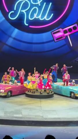 On a large stage, a group of approximately fifteen performers is arranged around two vintage convertible cars, one pink and one turquoise. In the center, several women in vibrant, polka-dotted dresses perform a dance, while others stand in or on the cars, striking poses with their arms raised or extended. The men wear suits in bright colors. Above them, a large backdrop displays the word "Roll" in neon-style script, accompanied by a stylized boombox graphic. The stage is illuminated with bright, colorful lighting, and a few audience members can be seen in the foreground.
