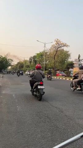 This is a vertical, eye-level shot of a street scene with moderate traffic. The road is paved and appears to be in fair condition, with some visible cracks and wear. Multiple motorcycles and a few cars are traveling in the same direction. The motorcycles are a mix of scooters and traditional-style bikes, most with one or two riders. Helmets are visible on many of the riders. The cars are modern SUVs. Trees line the right side of the road, with some foliage and a yellow flowering tree visible. Streetlights are present. The sky is a light, hazy orange, suggesting it might be early morning or late afternoon. Smoke is visible in the distance on the left side of the frame. The overall impression is a busy urban or suburban street during daylight hours.