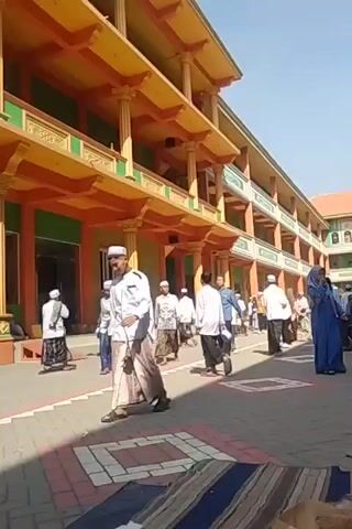 The video shows an outdoor scene with a multi-story building with orange and green accents. Many people, mostly men and some women and children, are present. Some are walking, while others are sitting on mats on the ground. Many of the men are wearing traditional Islamic attire, including kufis and sarongs. One man in the foreground is riding a hoverboard. The ground is paved with interlocking bricks. The overall atmosphere appears to be a gathering or event.