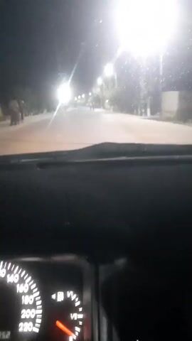 The video shows a view from inside a vehicle at night. The dashboard and speedometer are visible in the foreground, showing the needle at approximately 100. The road ahead is illuminated by streetlights. Several figures, appearing to be people, are walking or standing along the side of the road in the distance. Some trees and buildings are also visible. The overall scene depicts a nighttime drive through a populated area.