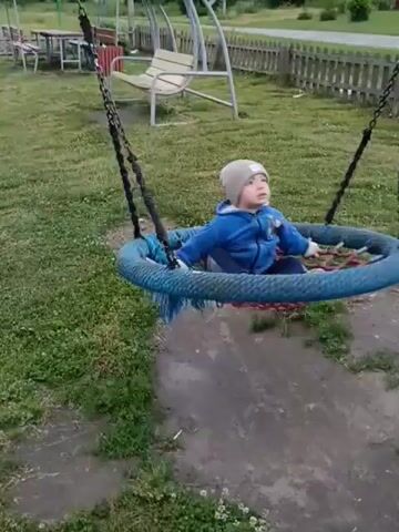 In Smolino, Russia, a young child, wearing a light brown beanie and a blue jacket, is enjoying a swing in a grassy outdoor area. The swing is a round, netted seat with a thick blue rim and red netting, suspended by thick black chains. The child appears to be seated comfortably, looking forward with a neutral expression.

The setting suggests a rural or suburban environment. In the background, there is a wooden fence, a paved path, and a large, multi-colored swing set with a bench seat. Beyond the fence, several houses with red and green roofs are visible, surrounded by trees and greenery. The overall impression is a bright, daytime scene, likely in the late spring or summer, with overcast skies. The child is the sole focus of the video, engaged in the simple activity of swinging.