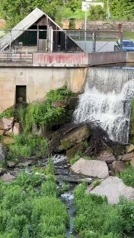 The video shows a multi-tiered waterfall cascading over rocks and concrete structures. A small, weathered wooden building with a pitched roof is situated above the upper part of the waterfall. Lush green vegetation grows around the waterfall and on the surrounding rocks. A concrete wall with an opening and some stairs is visible to the left of the waterfall. In the background, a road with a car is visible, as are trees and a distant building. The water appears to be flowing vigorously.