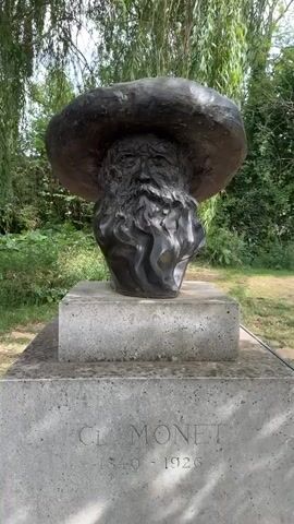 A bronze bust of Claude Monet, depicting him with a full beard and a wide-brimmed hat, stands on a concrete pedestal. The pedestal is inscribed with "CL MONET" and the years "1840 - 1926." The bust is situated outdoors, with lush green foliage and trees visible in the background. The lighting suggests it is daytime. The camera pans slightly to the left and then right, offering different angles of the sculpture.