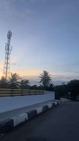 This video captures a serene moment at dusk in Banda Aceh, Indonesia. The scene is dominated by a vast, pale blue sky, gradually transitioning into soft hues of yellow and orange near the horizon, indicative of a sunset. Scattered, wispy clouds add texture to the sky.

In the foreground, a white railing with a golden accent runs horizontally, suggesting a viewpoint from an elevated position, possibly a balcony or bridge. Below the railing, a dark line of trees, including several prominent palm trees with their distinctive fronds, forms a silhouetted backdrop. The dense foliage hints at a tropical environment.

A thin, dark line in the sky, likely a distant airplane, is visible in the upper part of the frame, adding a touch of movement to the static scene. Utility poles and wires are also subtly present, interwoven with the tree line, signifying the presence of infrastructure. The overall mood is peaceful and tranquil, evoking the quiet beauty of the day's end. The lighting suggests it is twilight, with the sun having recently set or about to set. There is no visible text in the media file.