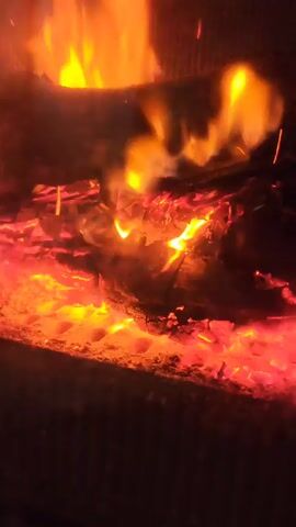 The video shows a close-up view of a fire burning in a fireplace or stove. Logs are visible at the top with flames emanating upwards. Below, there are glowing embers and coals, casting a warm, red light. There is some smoke or haze, possibly from the fire itself. The background is dark and out of focus.