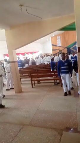 The video shows a group of people inside what appears to be a church or large hall. Most of the individuals are wearing white uniforms with some wearing blue sweaters over their white shirts. They are standing and moving around. The interior of the building has rows of wooden pews. There is a stage-like area at the far end with decorations. The camera appears to be held by someone who is walking past the group. The lighting in the room is moderately bright.