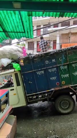 The video shows a side view of a garbage truck parked on a street. A person wearing a plaid shirt is standing on the back of the truck, appearing to adjust some of the cargo, which includes large white bags. The truck has blue and green sides with text in an Indian script. There are buildings with balconies and power lines in the background, suggesting an urban or semi-urban environment. The ground appears wet, possibly from rain. A green corrugated roof extends over the foreground.