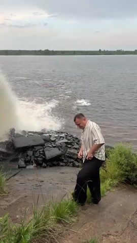 A man in a striped shirt and dark pants stands on a concrete surface near a large, broken concrete structure where water is gushing out. The structure appears to be part of a dam or levee, with large chunks of concrete debris scattered around its base. The water is creating a powerful surge and waves as it flows into a body of water that extends to the horizon. The man is looking down and slightly to his right, with his left hand near his waist. The sky is overcast, and there is green vegetation along the edge of the concrete surface.