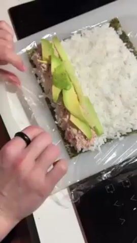 A pair of hands is shown in this close-up shot rolling a sushi roll. The hands, adorned with light pink nail polish and a black ring, are carefully folding the nori seaweed sheet filled with white rice, tuna, and sliced avocado. The roll is being wrapped in plastic wrap, and the hands are using a gentle but firm pressure to seal the roll. The surface beneath is a white cutting board, with a black stovetop visible in the background. The overall scene depicts the process of making sushi at home.